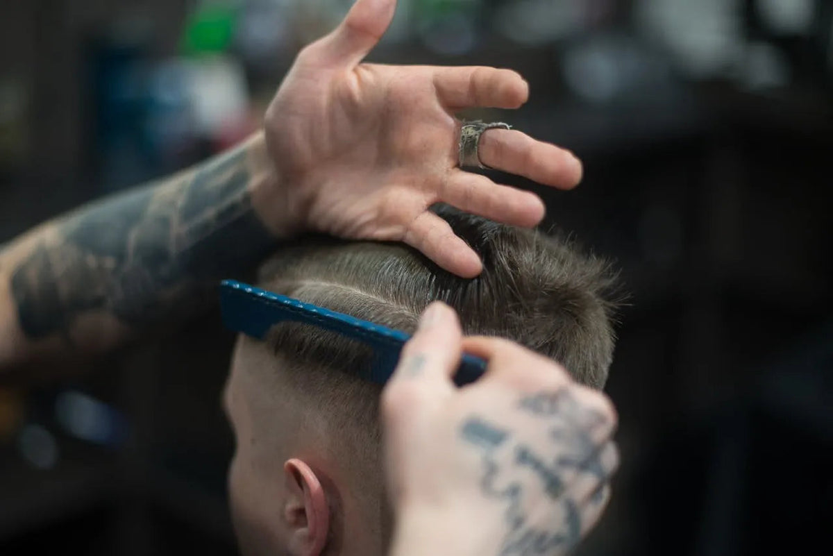 Friseør med tatoveringer stiler hår med blå kam i barberbutikk